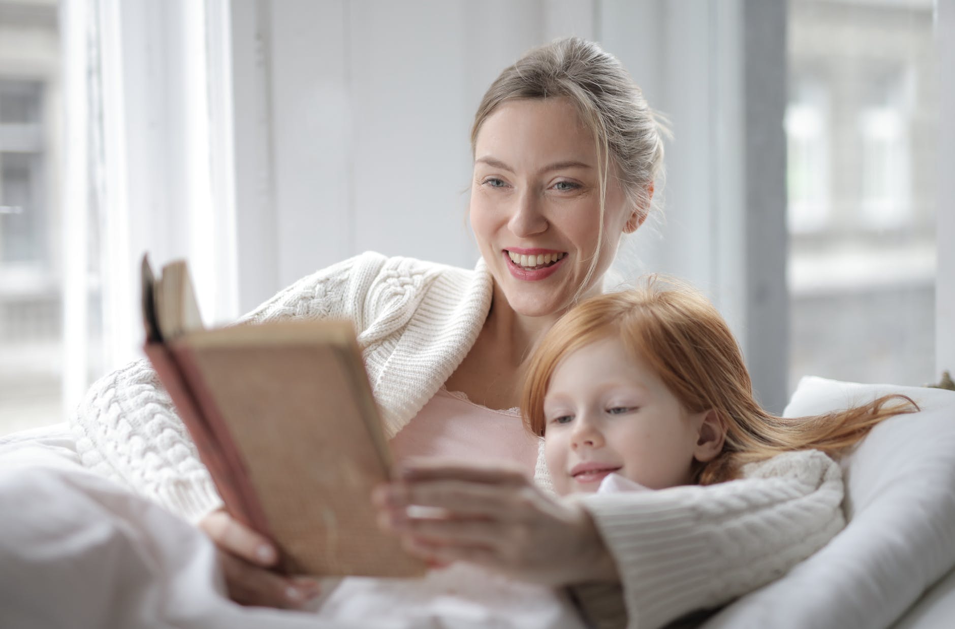 woman holding book lying on bed beside girl