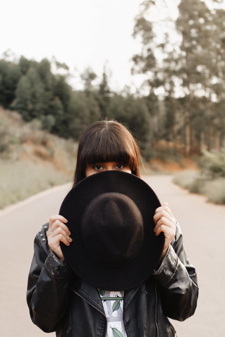 selective focus photo of woman standing in the middle of a road while covering part of her face with a fedora